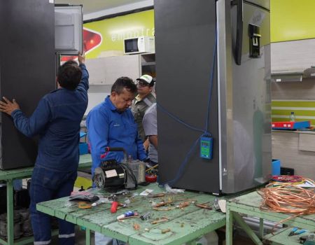 A man is repairing a fridge. Some men are behind him, and tools are over the table.